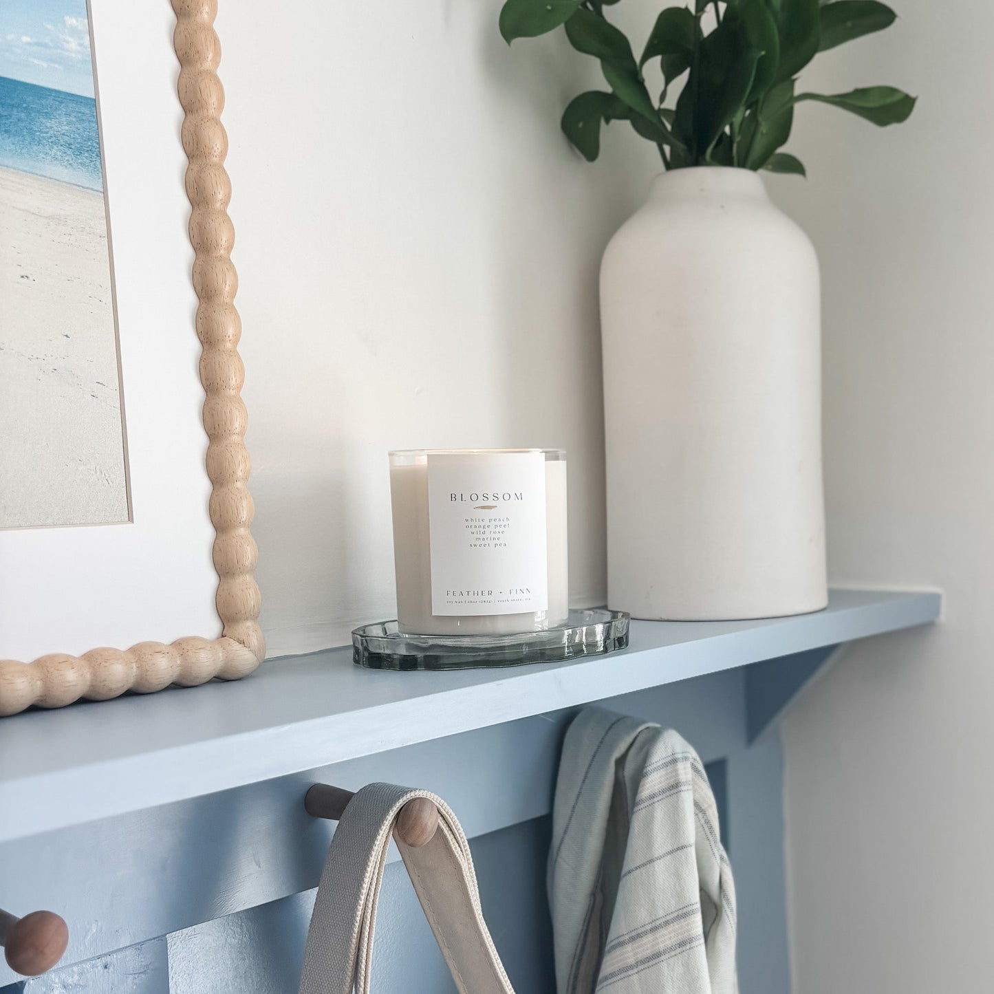 blossom candle on shelf with picture frame and white vase filled with greens