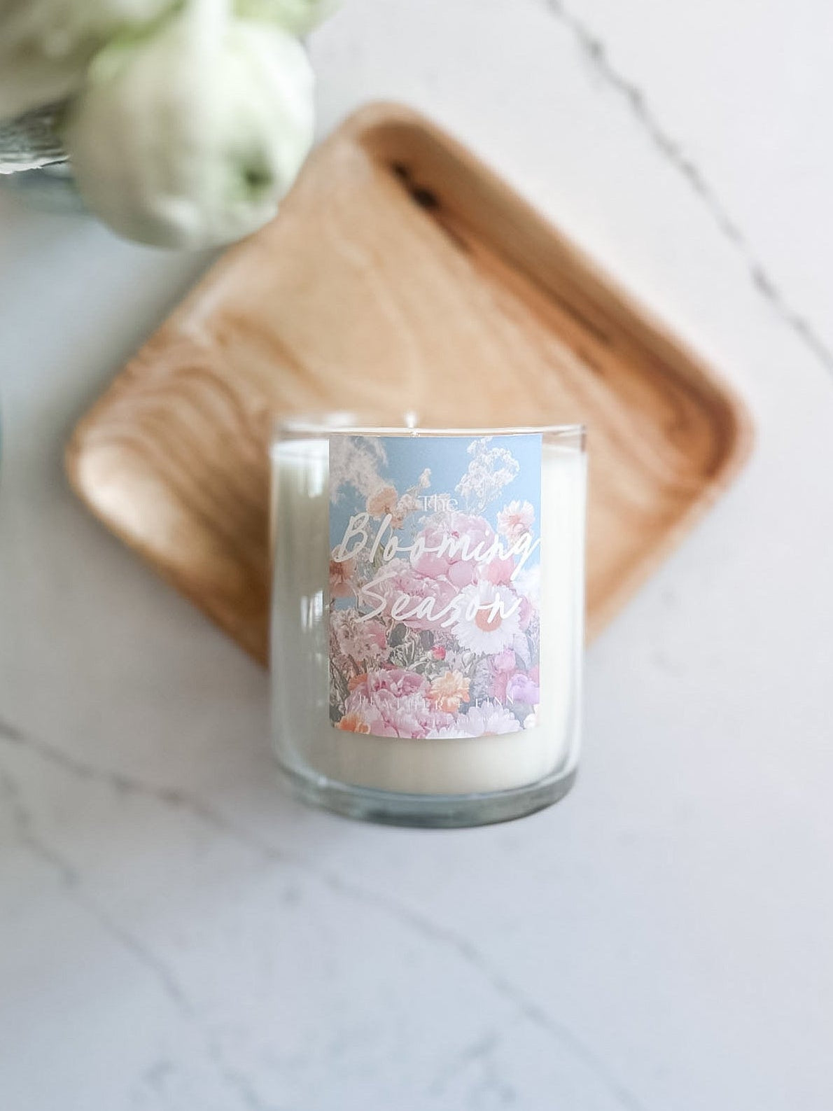 Candle with floral design and text on a marble surface with flowers and a wooden coaster.