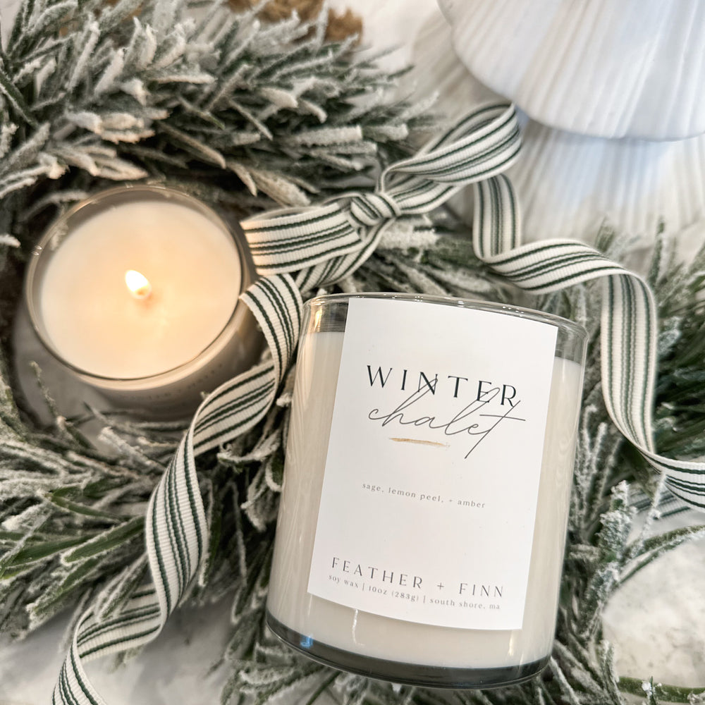 A lit candle alongside a candle labeled winter chalet tucked into winter greens.