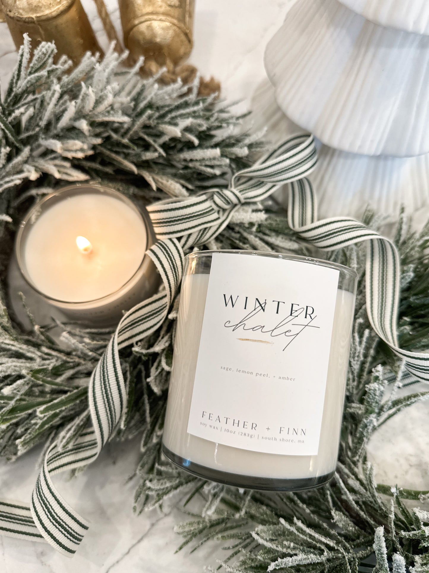 A lit candle alongside a candle labeled winter chalet tucked into winter greens.