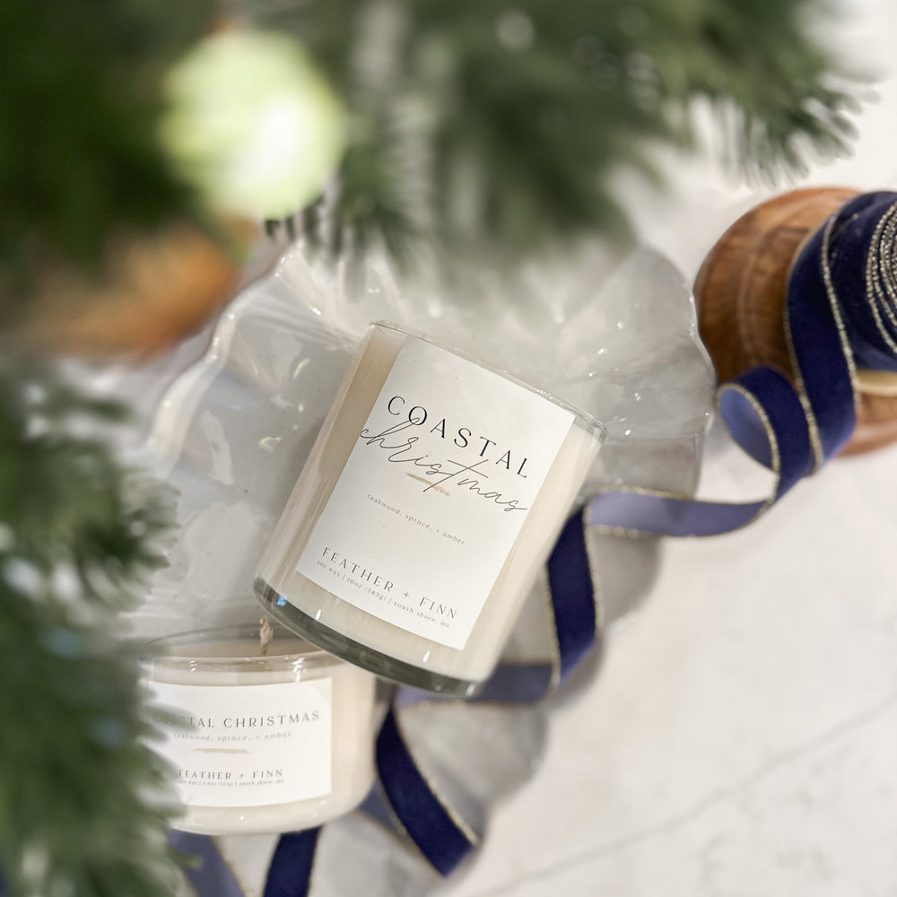 Candle labeled 'Coastal Christmas' with a blue ribbon under a Christmas tree.